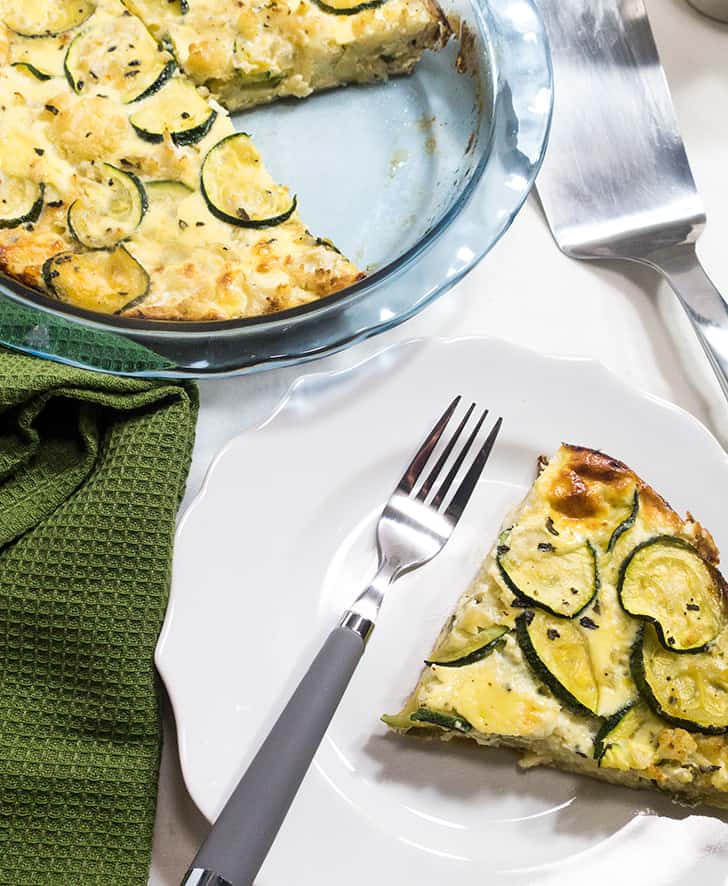  a slice of zucchini pie on a white plate with a grey fork. A green napkin beside the plate.