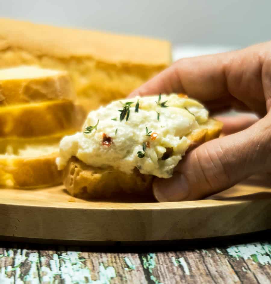 Baked ricotta on a slice of a baguette garnished with fresh thyme and red chili pepper flakes that's being grabbed with a hand.