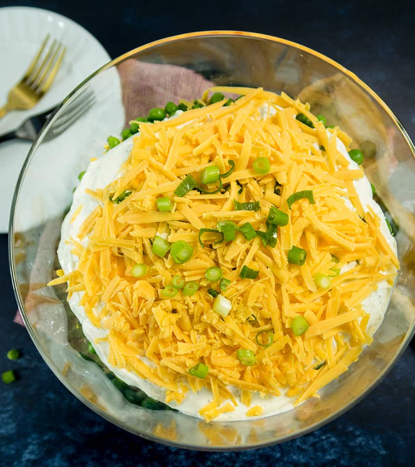 Simple Make Ahead 7 Layer Salad in a clear glass bowl looking down on the top with shredded cheddar cheese and  chopped green onions.