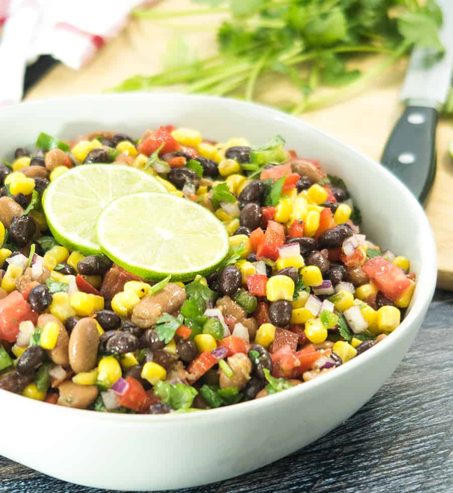 black bean corn salad in a white bowl with fresh chopped cilantro and lime slices. Cilantro and a knife in the background on a cutting board.