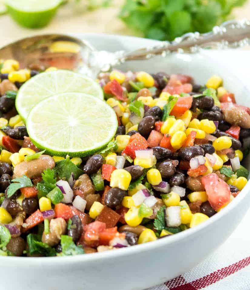 black bean corn salad in a white bowl with lime slices and a silver spoon for serving.