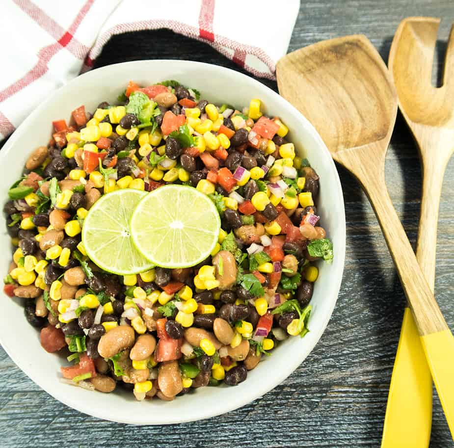 black bean corn salad in a white bowl with lime slices. Wooden salad spoons beside the bowl for serving