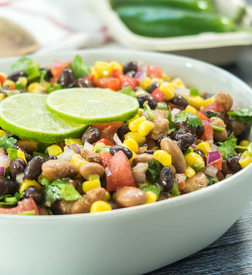 black bean corn salad in white bowl with lime slices and fresh chopped cilantro. Jalapeños in the background.