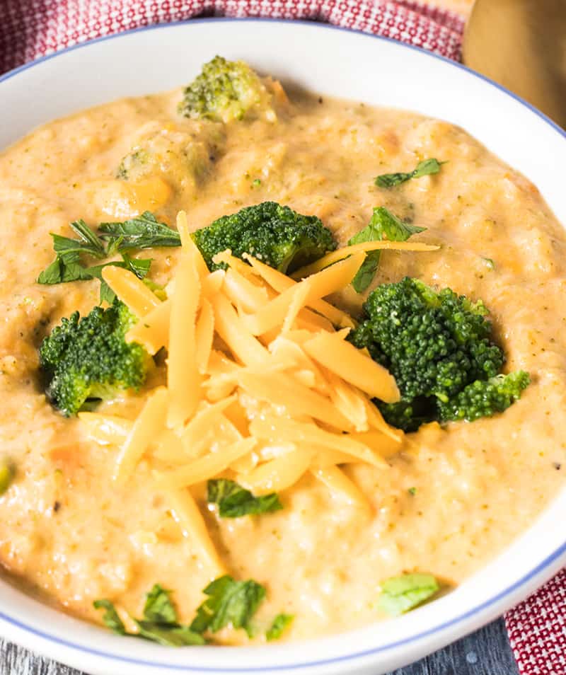 Crockpot Broccoli Cheese Soup in a white bowl with blue trim topped with extra shredded cheese and broccoli florets.