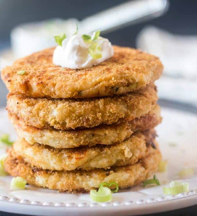 Mashed Potato Cakes stacked on a white plate topped with sour cream & green onions.