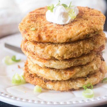 Mashed potato cakes stacked on a plate topped with sour cream and green onions.