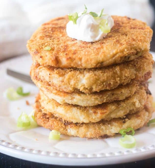 Mashed potato cakes stacked on a plate topped with sour cream and green onions.
