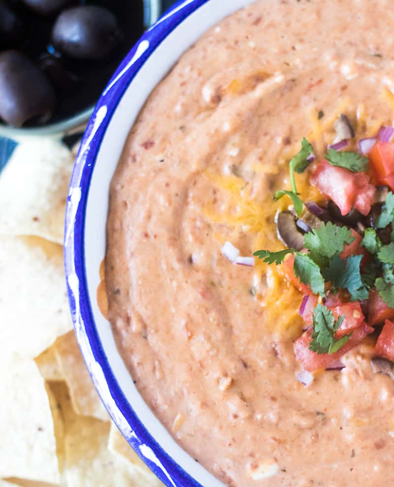 Bean dip in a bowl with chopped tomato, red onion, black olives and cilantro with tortilla chips beside it.