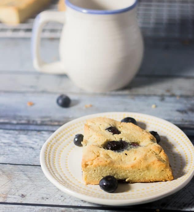 Blueberry Scone on a plate with some blueberries and coffee mug in background.