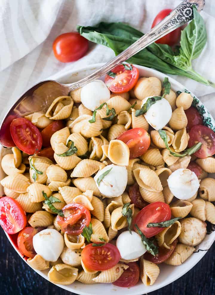 Caprese Pasta Salad in a bowl with a spoon and tomatoes and fresh basil in background.