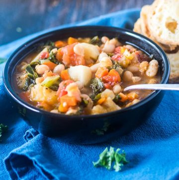 Bean stew loaded with an array of veggies including white beans, kale in a black bowl on a blue towel.