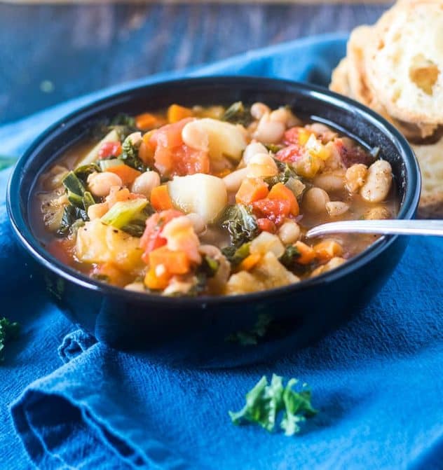 Bean stew loaded with an array of veggies including white beans, kale in a black bowl on a blue towel.