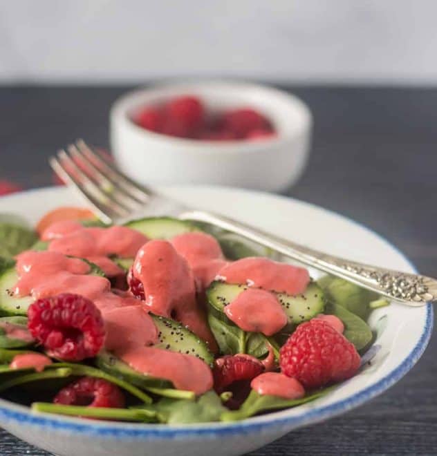 Spinach salad in a white bowl with cucumbers, raspberries and topped with raspberry vinaigrette and a bowl of raspberries in the background.