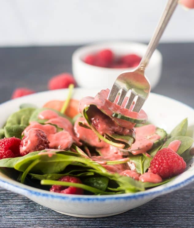 Spinach salad with raspberry vinaigrette on a silver fork with raspberries in a white bowl in the background.