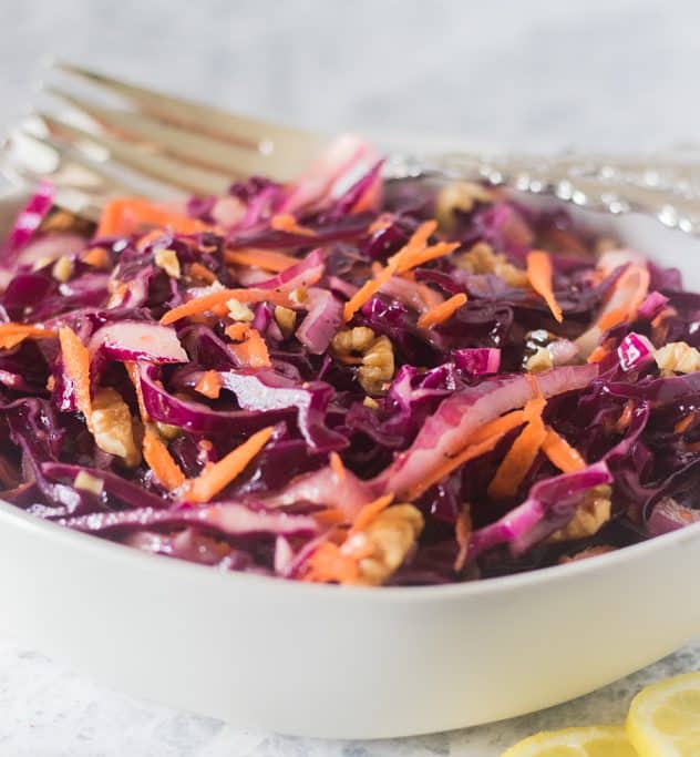 Red cabbage salad in a white bowl with silver serving spoons.
