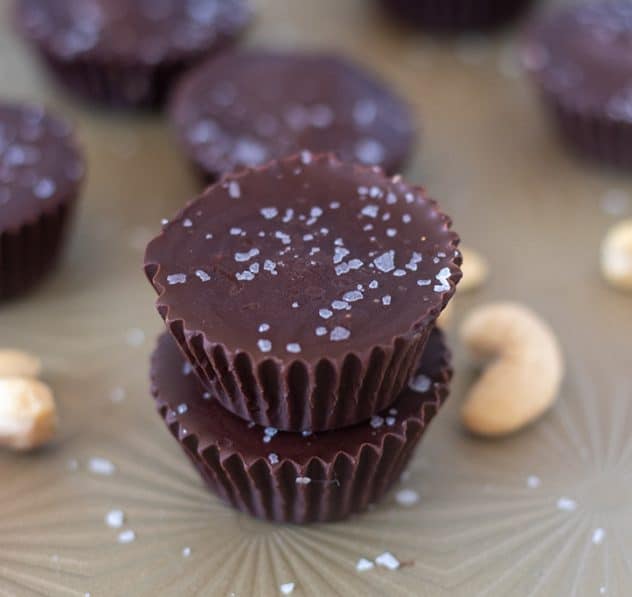 Dark Chocolate Cashew Nut Butter Cups stacked on baking try with cashews in background.