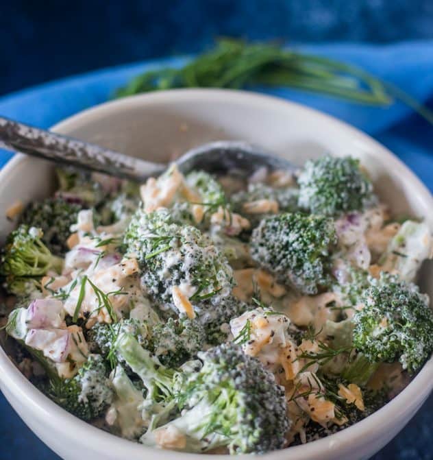 Broccoli Salad in a white bowl with a silver serving spoon with fresh dill and blue napkin in background.