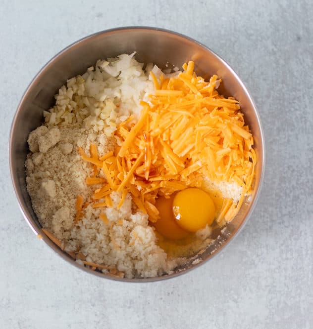 mixing bowl with riced cauliflower, shredded cheddar cheese, almond flour, eggs, chopped onion, garlic and hot sauce.