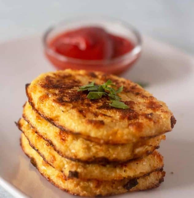 Cauliflower Hash Browns stacked on a plate with some ketchup in a bowl.