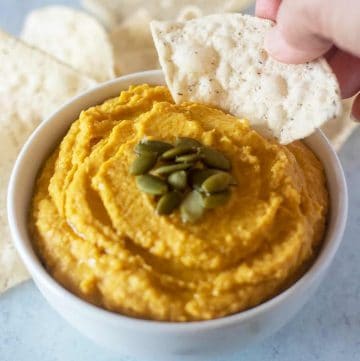 Pumpkin hummus in a white bowl being dipped with a tortilla chip.