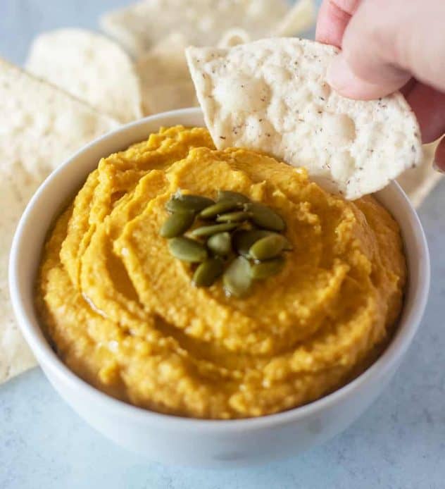 Pumpkin hummus in a white bowl being dipped with a tortilla chip.