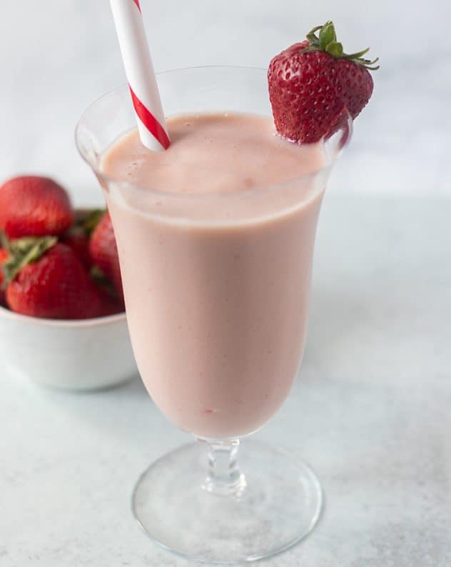 Strawberry milkshake in a glass with straw and garnished with a fresh strawberry. Bowl of strawberries in the background.