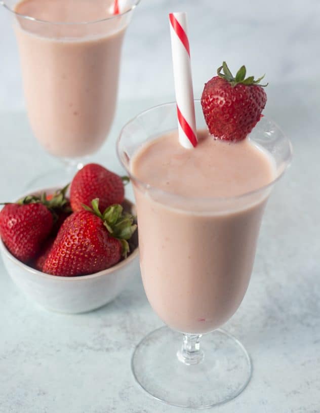 Strawberry milkshake in a glass with a straw and garnished with a fresh strawberry. and bowl of strawberries in background