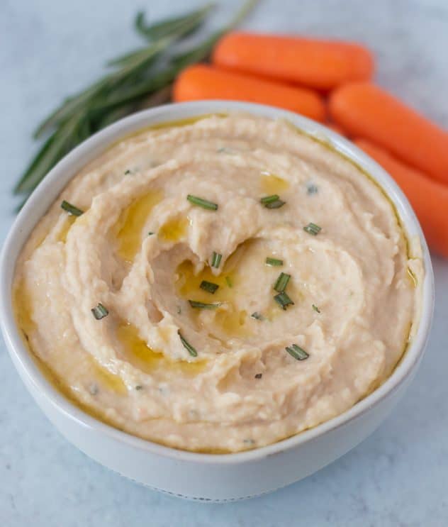 White bean dip in a bowl with some baby carrots and rosemary beside the bowl