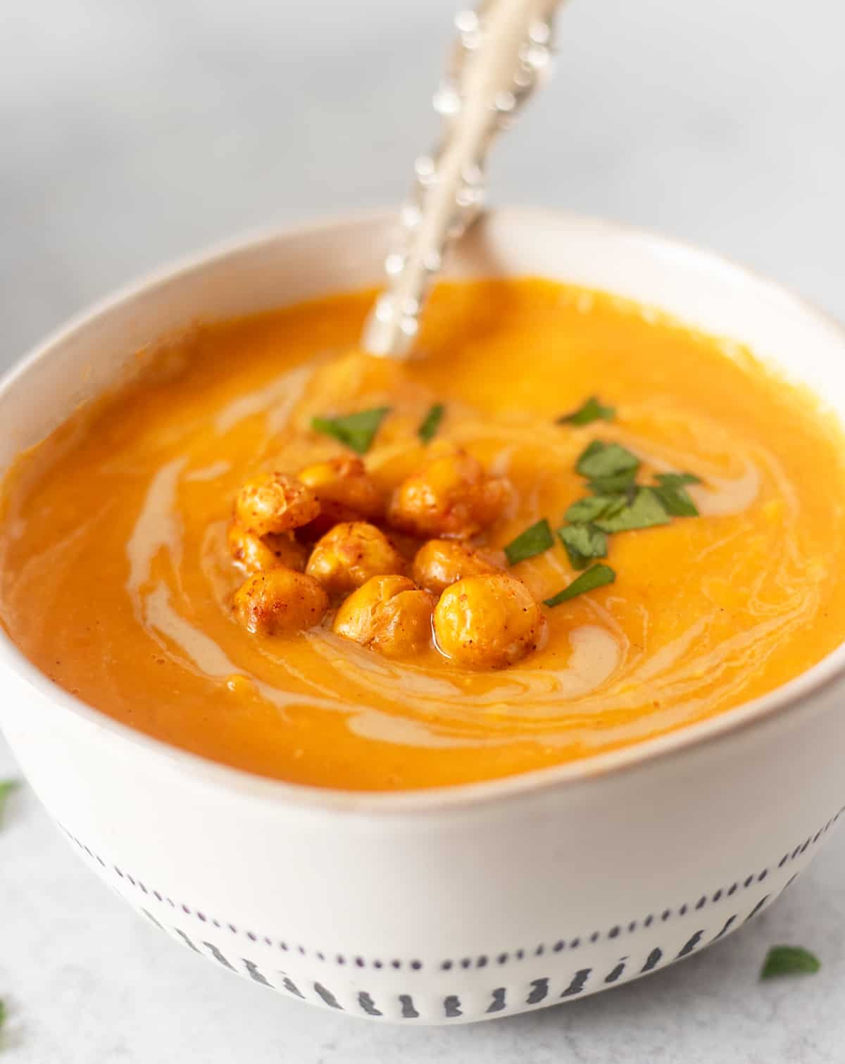sweet potato soup in a white bowl with a silver serving spoon and garnished with roasted chickpeas and chopped parsley