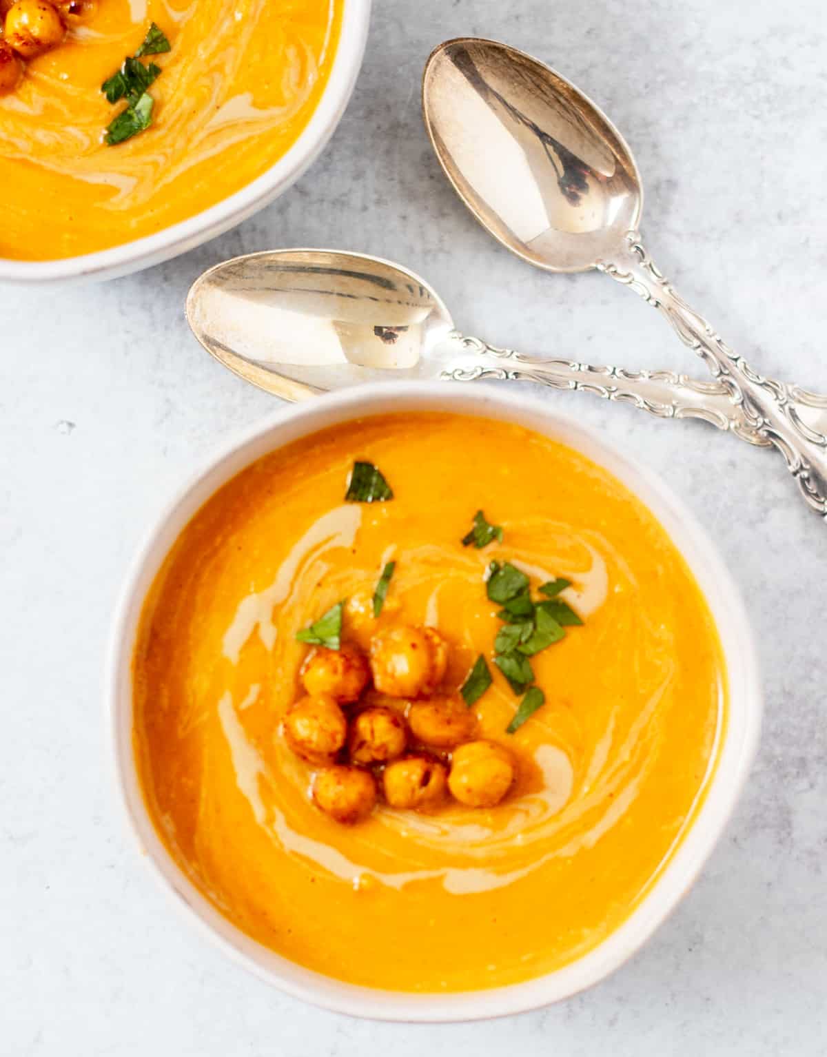 sweet potato soup in a white serving bowl topped with roasted chickpeas and parsley with silver serving spoons