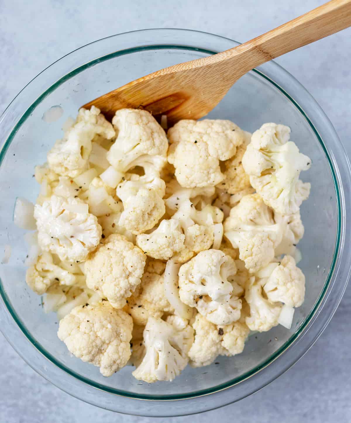 cauliflower florets in a mixing bowl with olive oil, salt & pepper and a wooden spoon