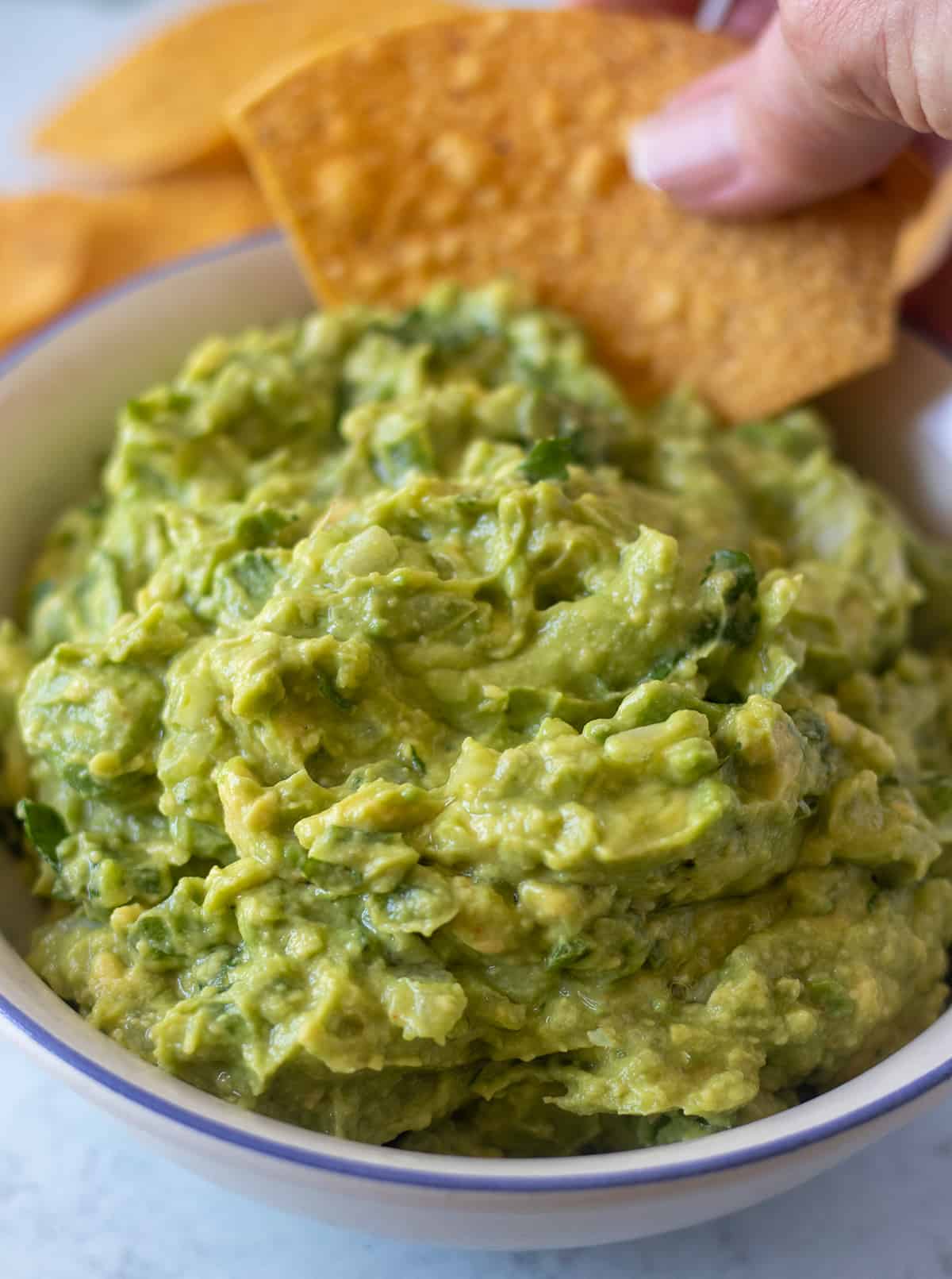 guacamole being in a white bowl with blue trim being scooped out with a tortilla chip