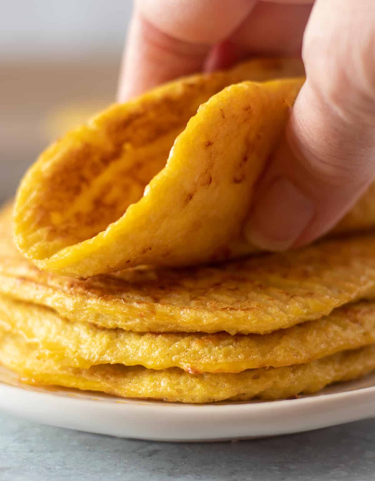 Cauliflower tortillas stacked on a plate with a hand grabbing and bending one.