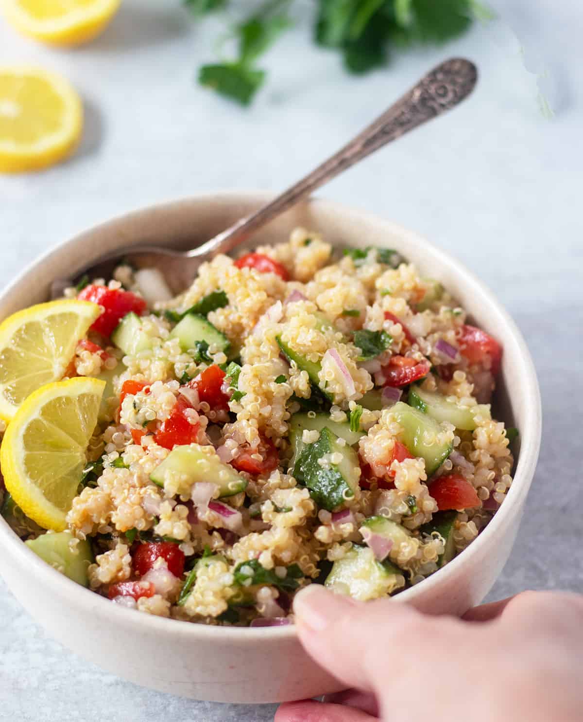 quinoa tabbouleh in a white bowl garnished with lemon slices and a silver spoon for serving. A hand holding the side of the bowl.