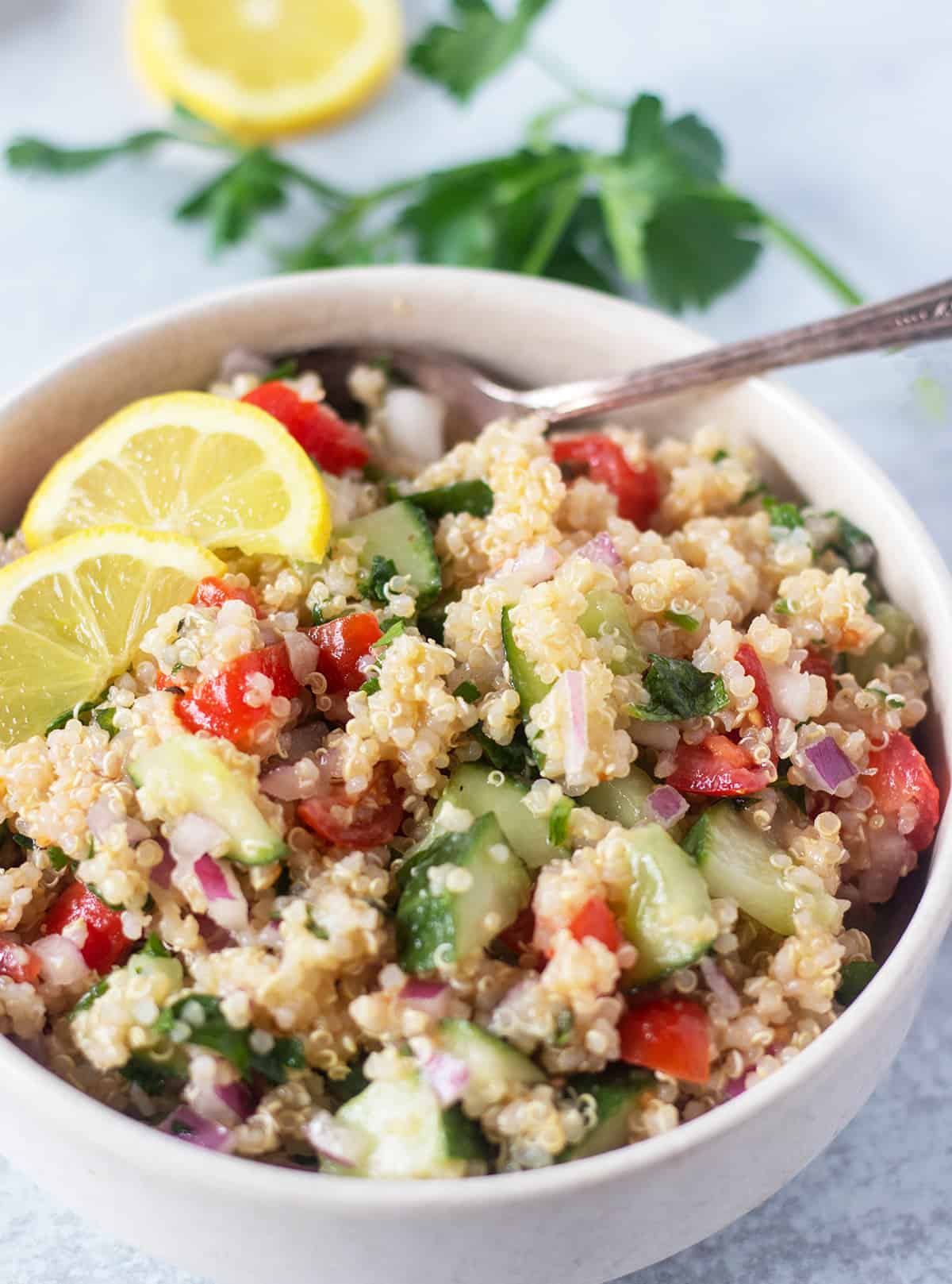 Quinoa tabbouleh in a white bowl with a silver spoon and garnished with lemon slices.