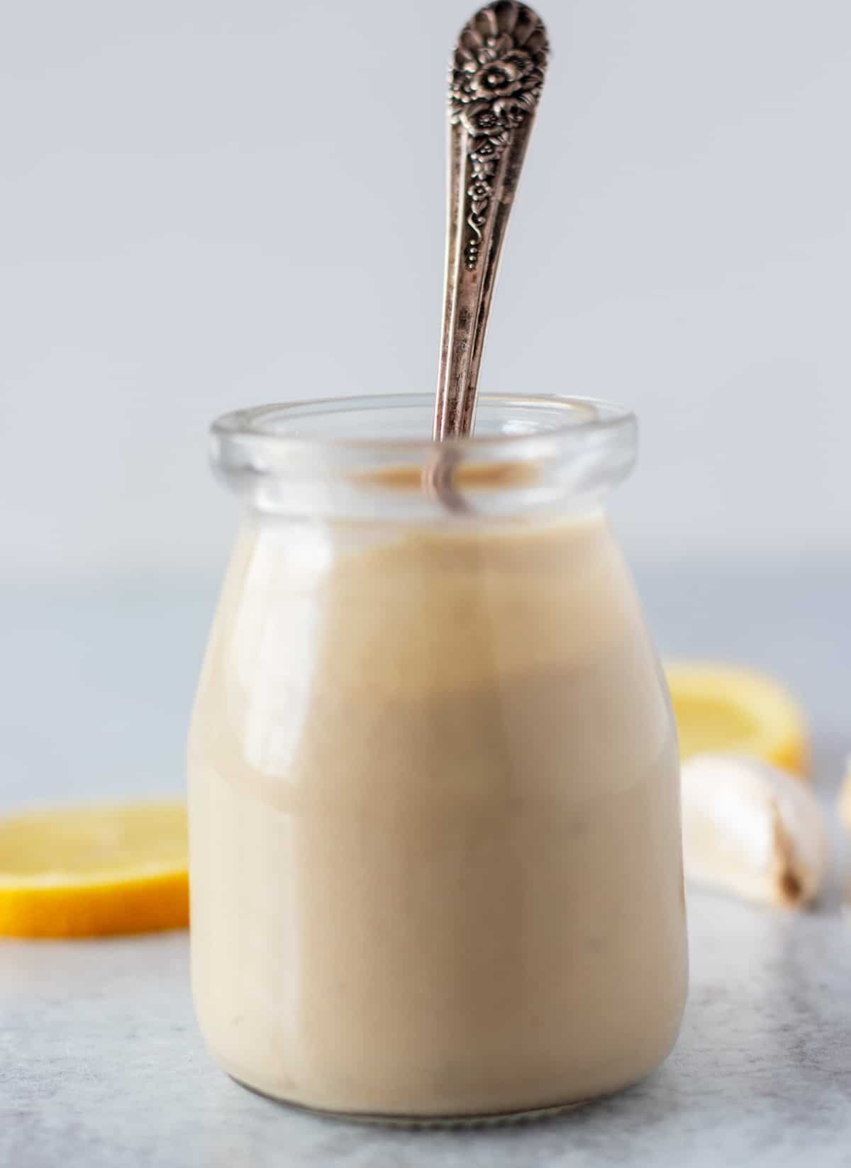 tahini dressing in a glass jar with lemon slices and garlic cloves in the background. a silver spoon in the jar for serving.