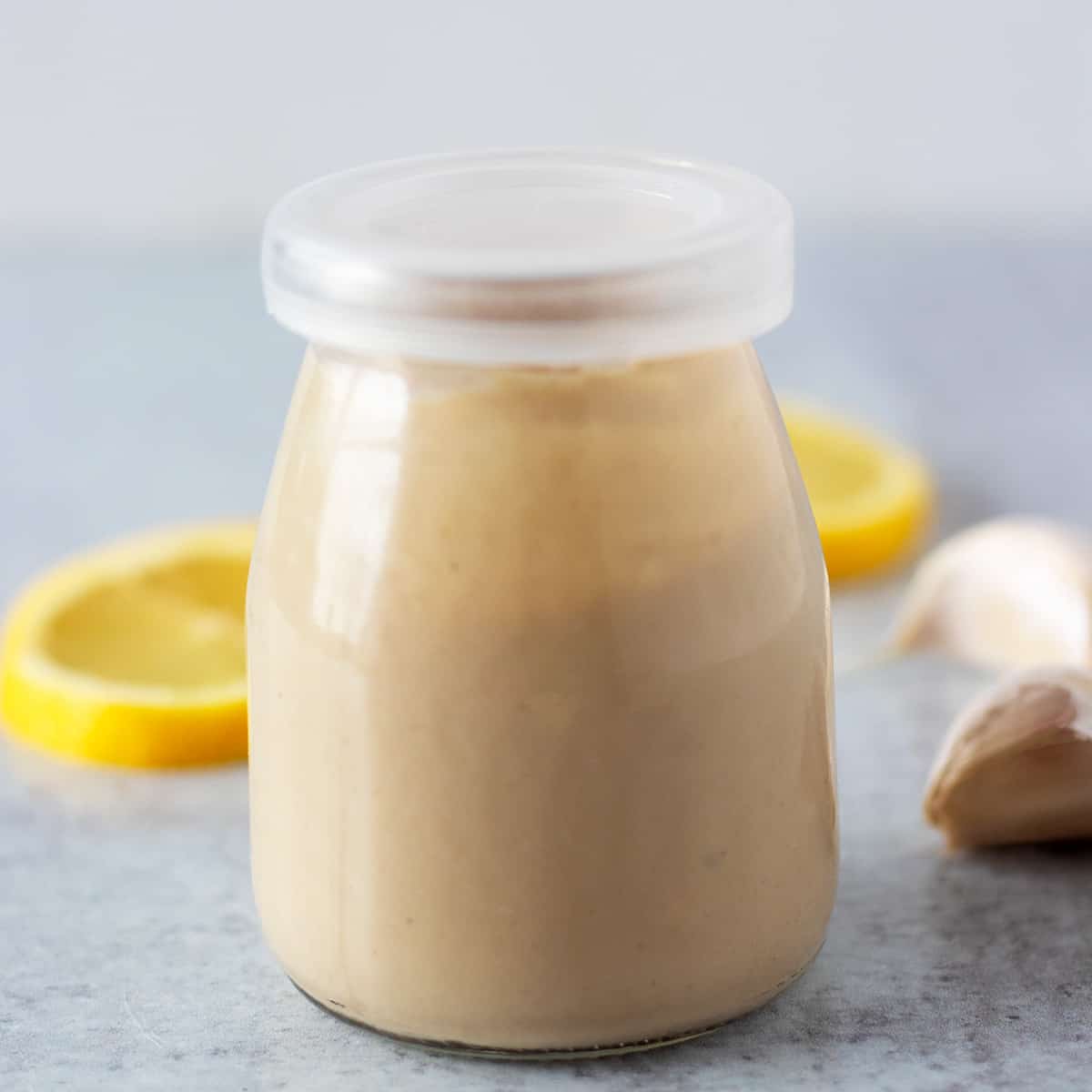 tahini dressing in a glass gar with lemon slices and garlic cloves in the background.