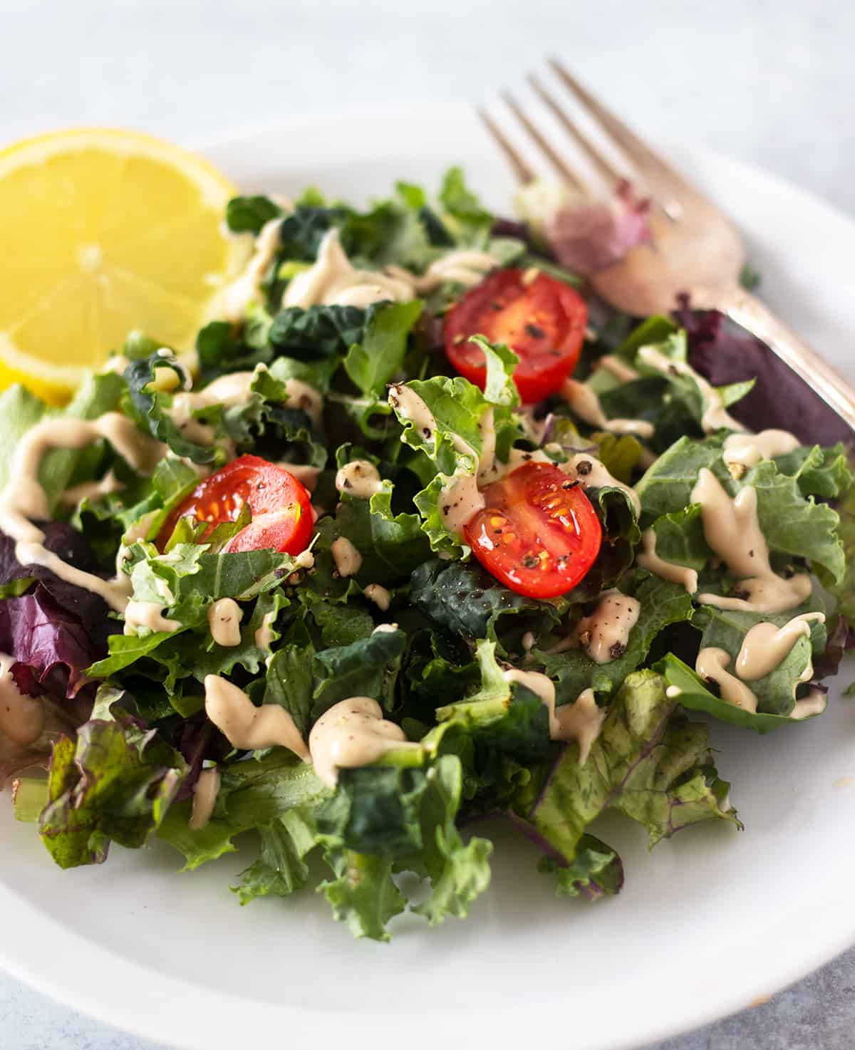 tahini dressing drizzled over a green salad with sliced grape tomatoes. A lemon wedge on side of the white plate and a silver fork for serving.