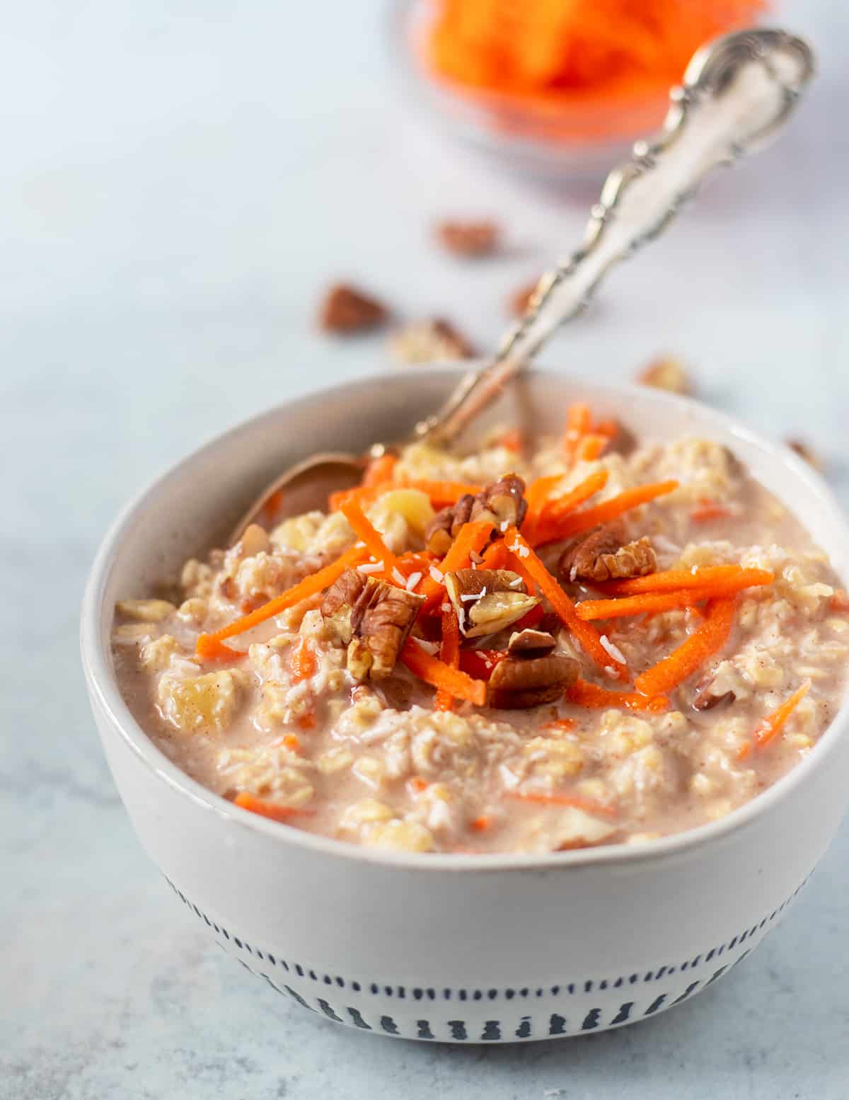 carrot cake overnight oats in a white bowl with blue trim that's garnished with shredded carrots, coconut flakes and chopped pecans. A silver spoon in the bowl for serving