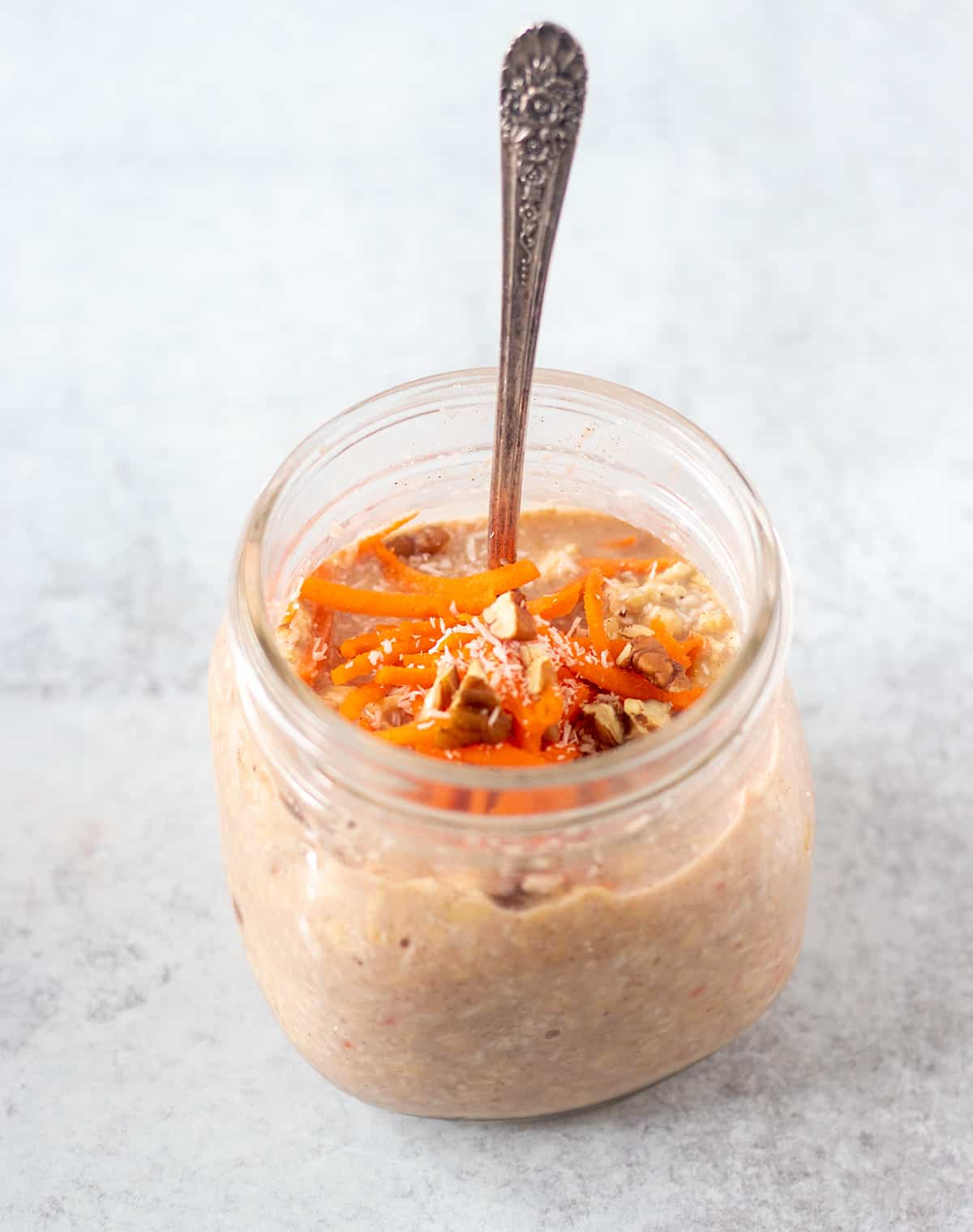 carrot cake overnight oats in a glass jar with a silver spoon. Topped with extra shredded carrots, chopped pecans, and shredded coconut