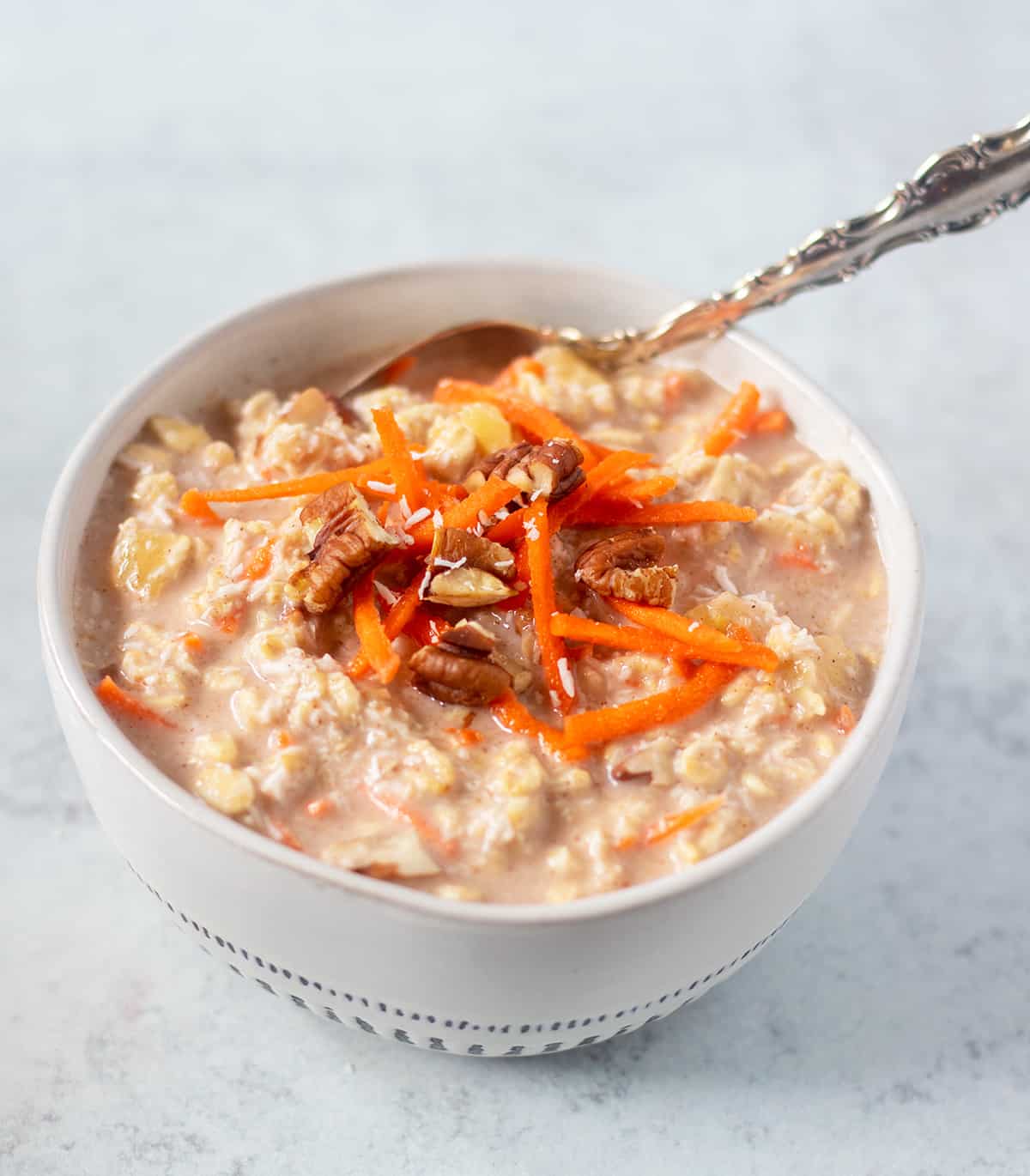 carrot cake overnight oats in a white bowl with blue trim topped with shredded carrots, chopped pecans and shredded coconut.