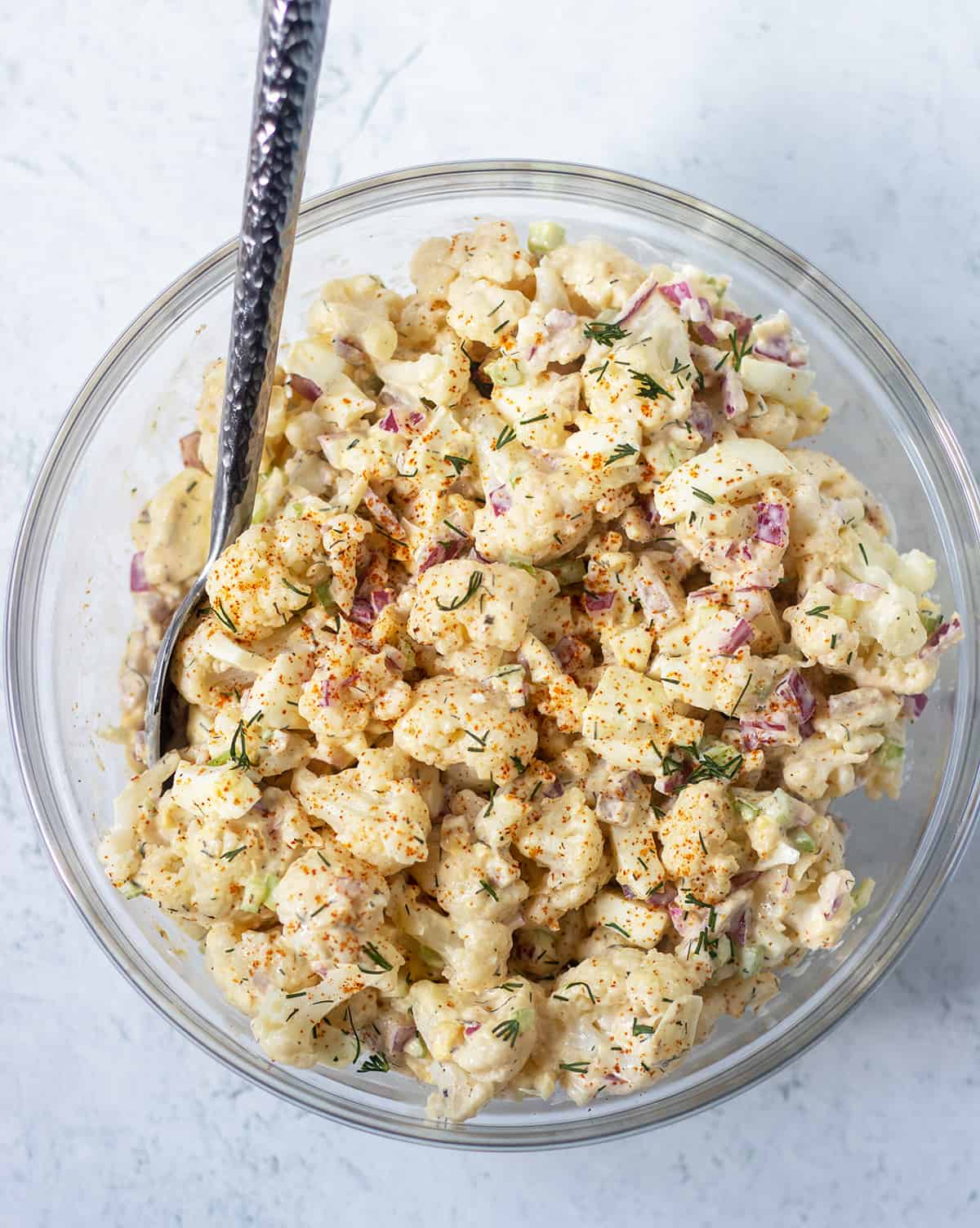 cauliflower potato salad in a clear glass bowl topped with fresh dill. A silver spoon in the bowl for serving.
