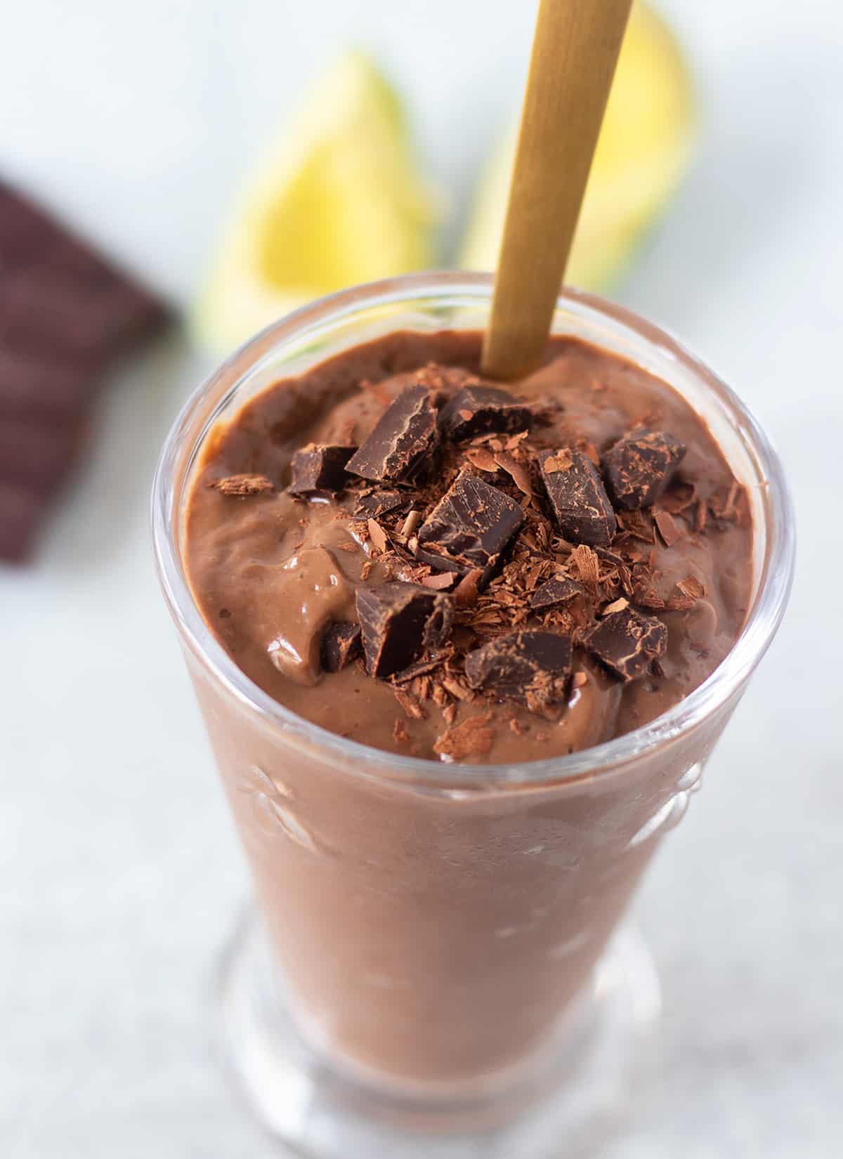 chocolate avocado smoothie garnished with extra shaved chocolate and chocolate chunks in a clear glass. A gold spoon in the glass for serving. 