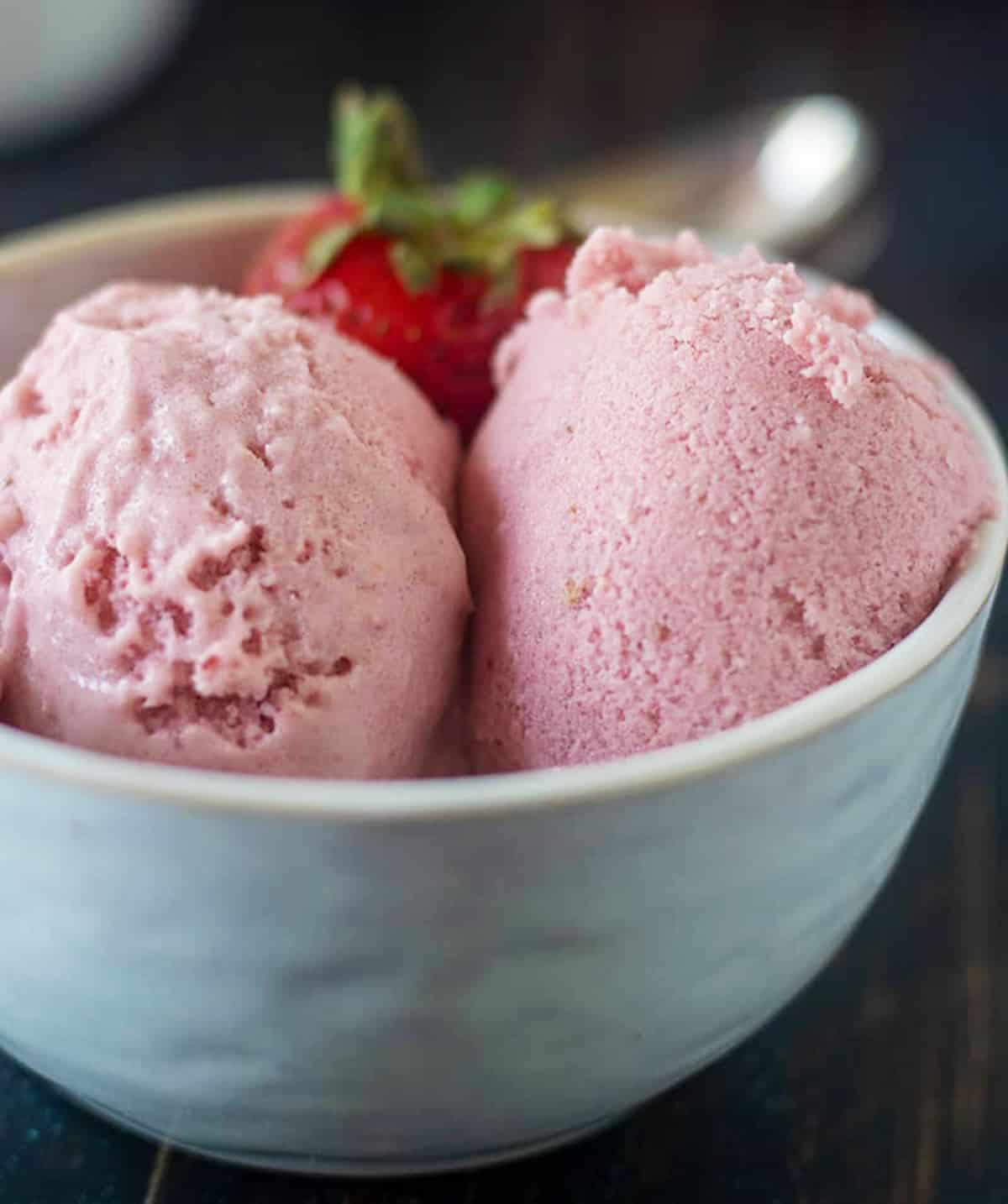 homemade strawberry ice cream in a white bowl topped with fresh strawberries. two silver spoons in the background.