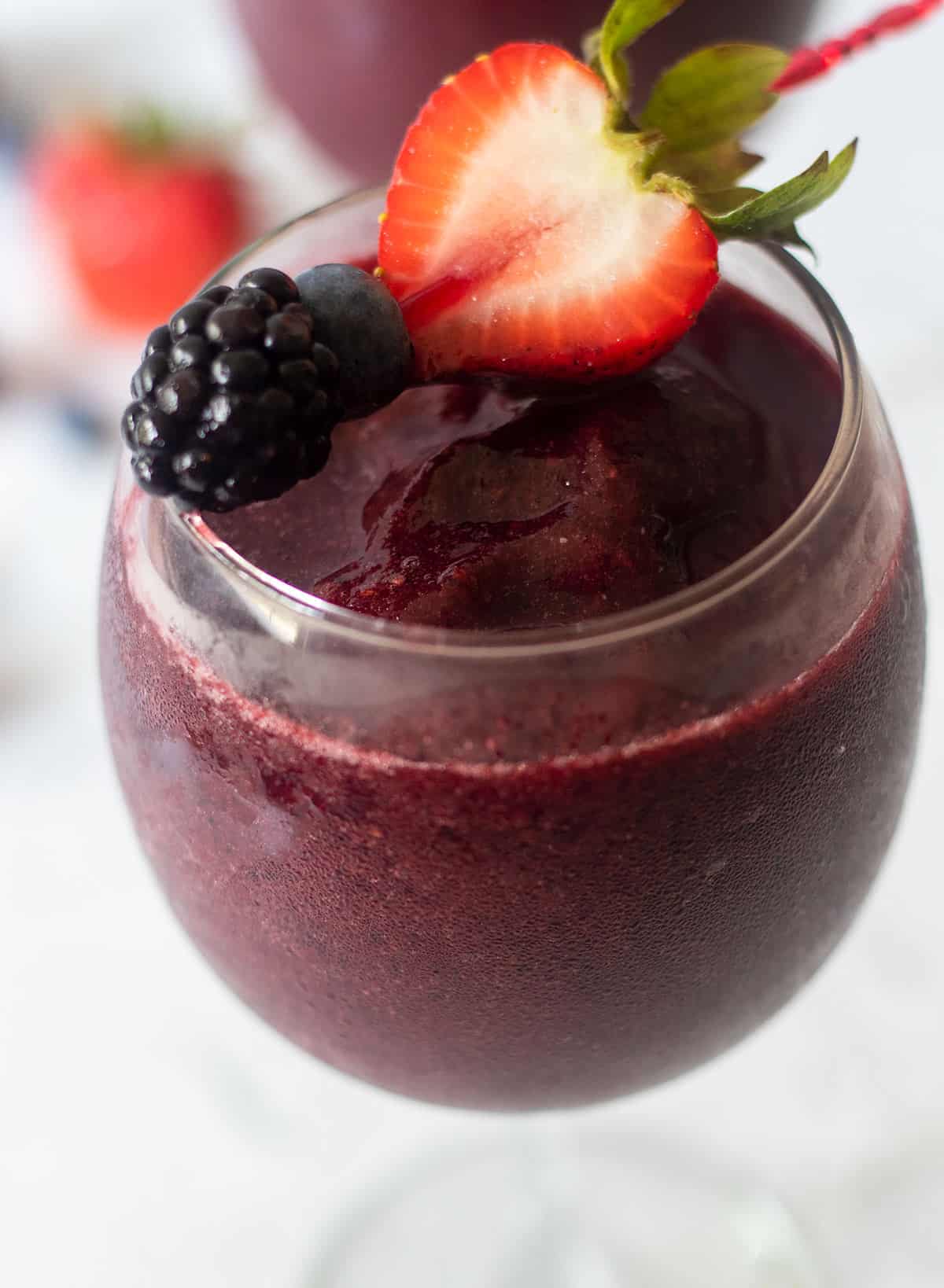 berry wine slushie in a clear wine glass topped with a toothpick that has a strawberry half, blueberry and blackberry on it.