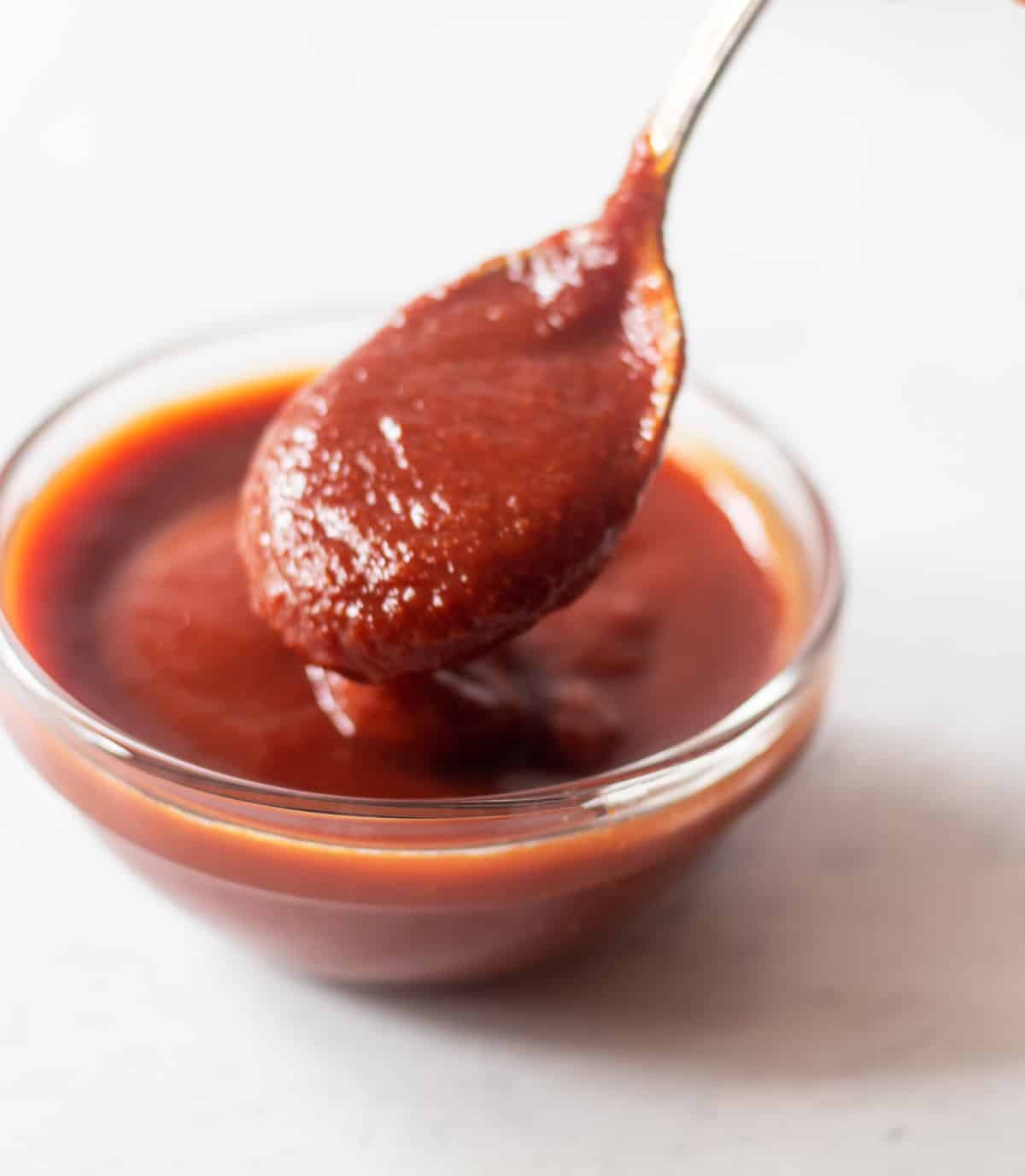 Homemade barbecue sauce in a clear glass bowl with a silver spoon scooping some out.