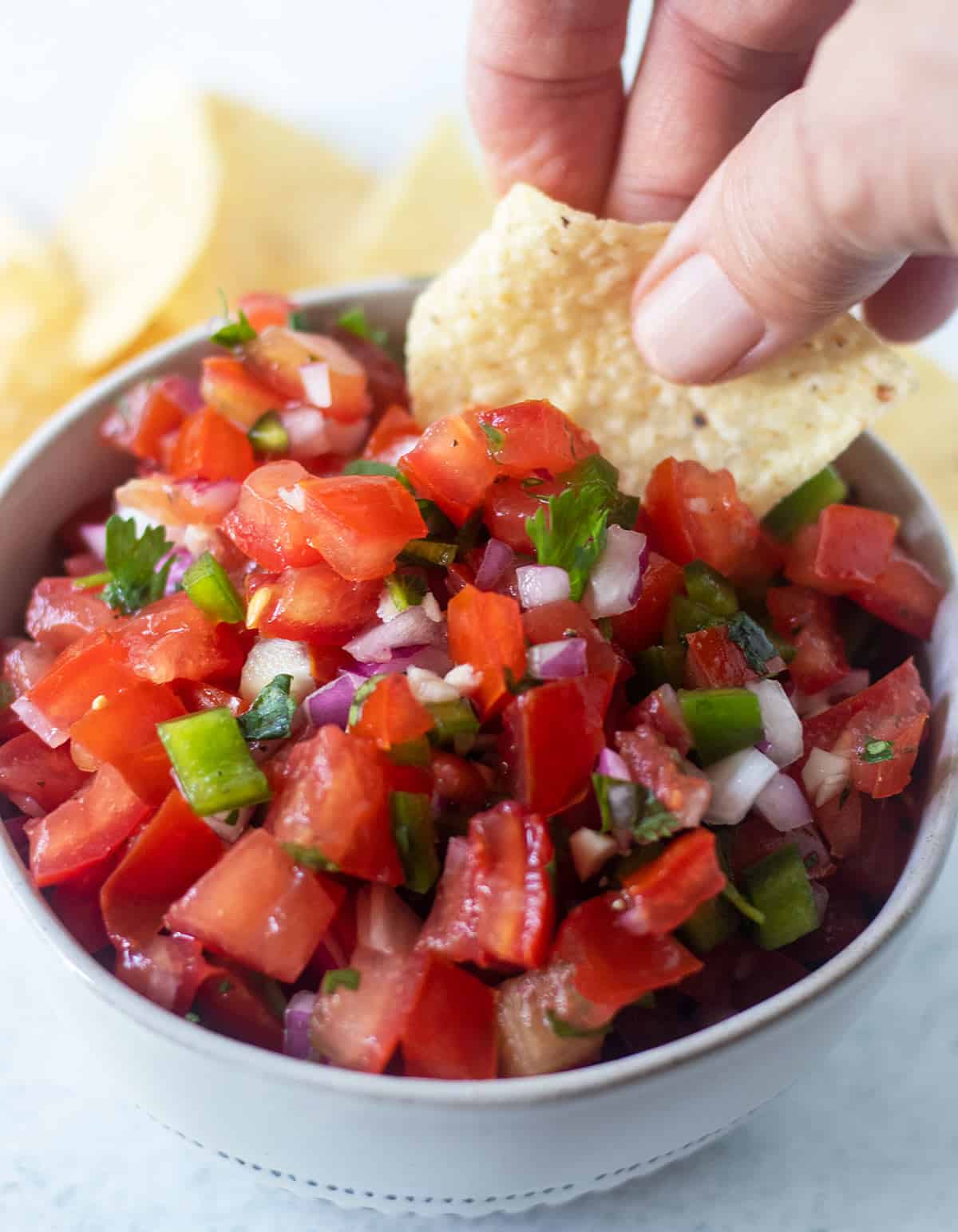 homemade pico de gallo in a white bowl with blue trim and a hand dipping some out with a tortilla chip.