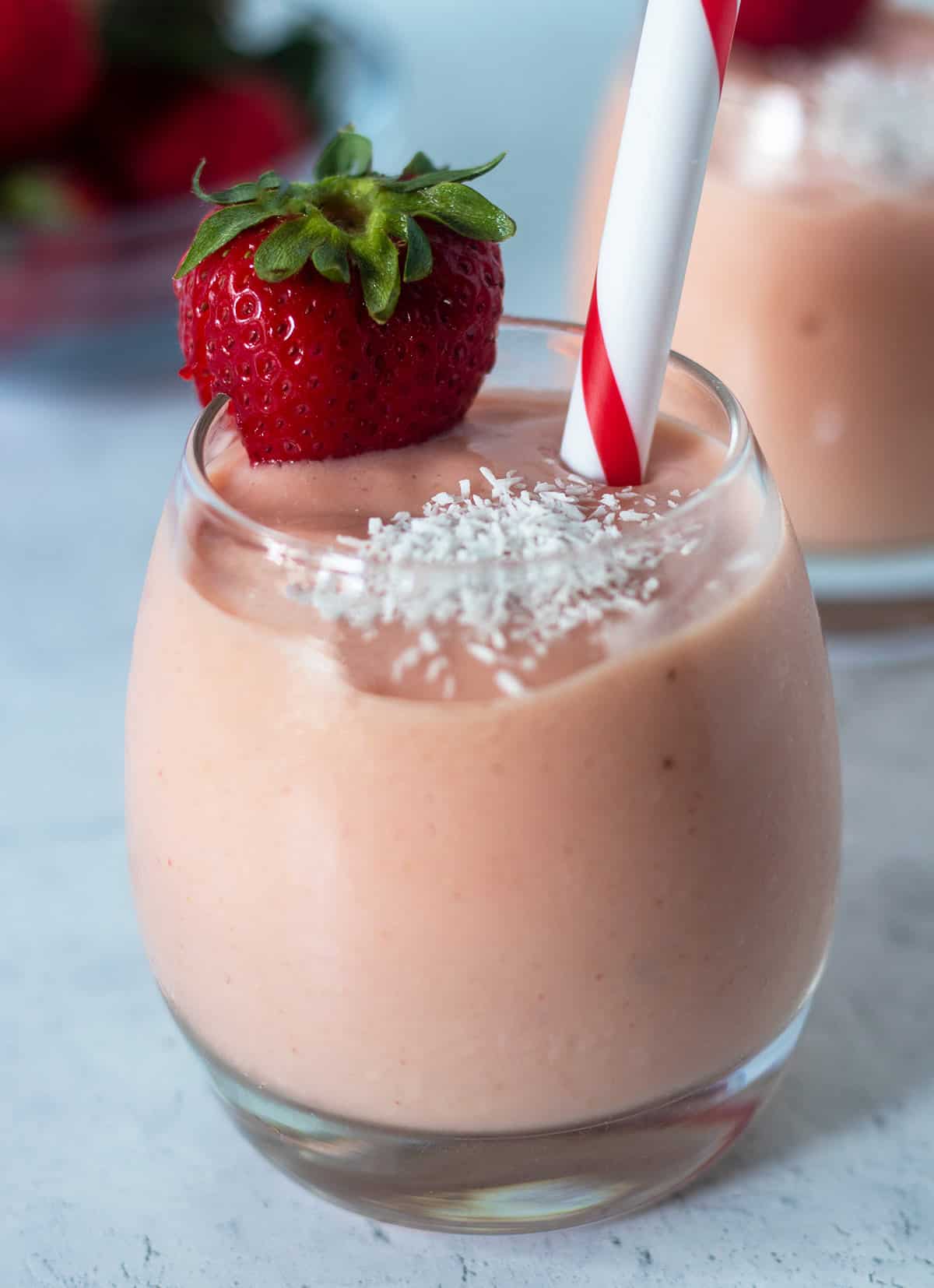 tropical smoothie in a clear glass garnished with a fresh strawberry on the side of the glass and shredded coconut on the smoothie. A red and white straw in the glass for serving.