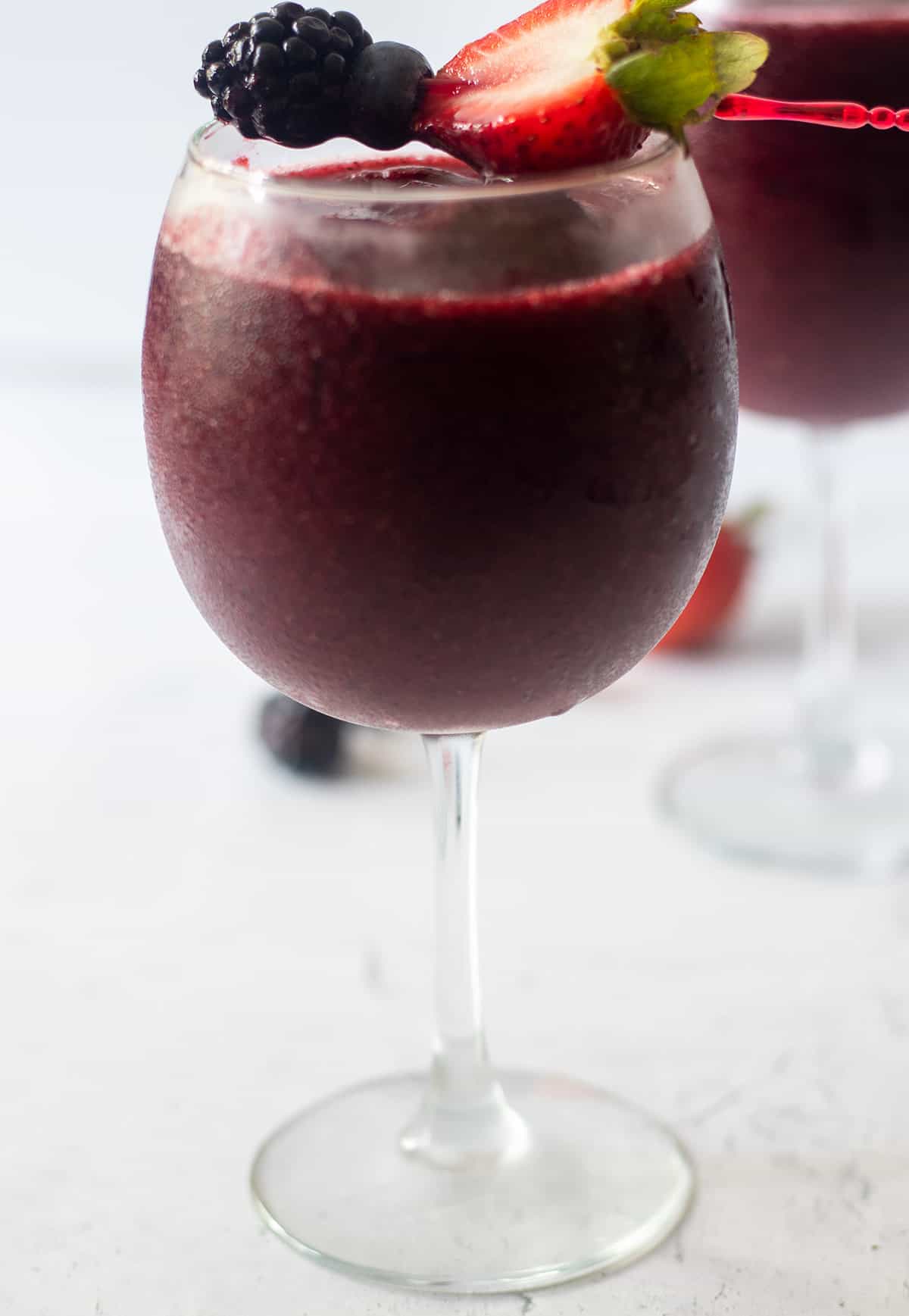 berry wine slushie in a clear wine glass with a toothpick with a half of a fresh strawberry, fresh blackberry and fresh blueberry on it.