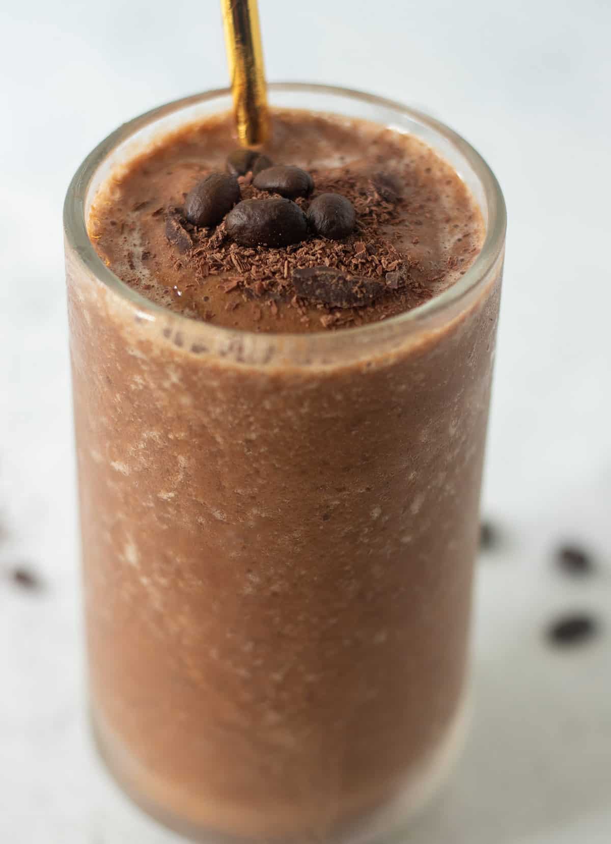 coffee smoothie recipe served in a clear glass with a gold straw. It's topped with dark shaved chocolate and fresh coffee beans. A gold straw in the glass for drinking.